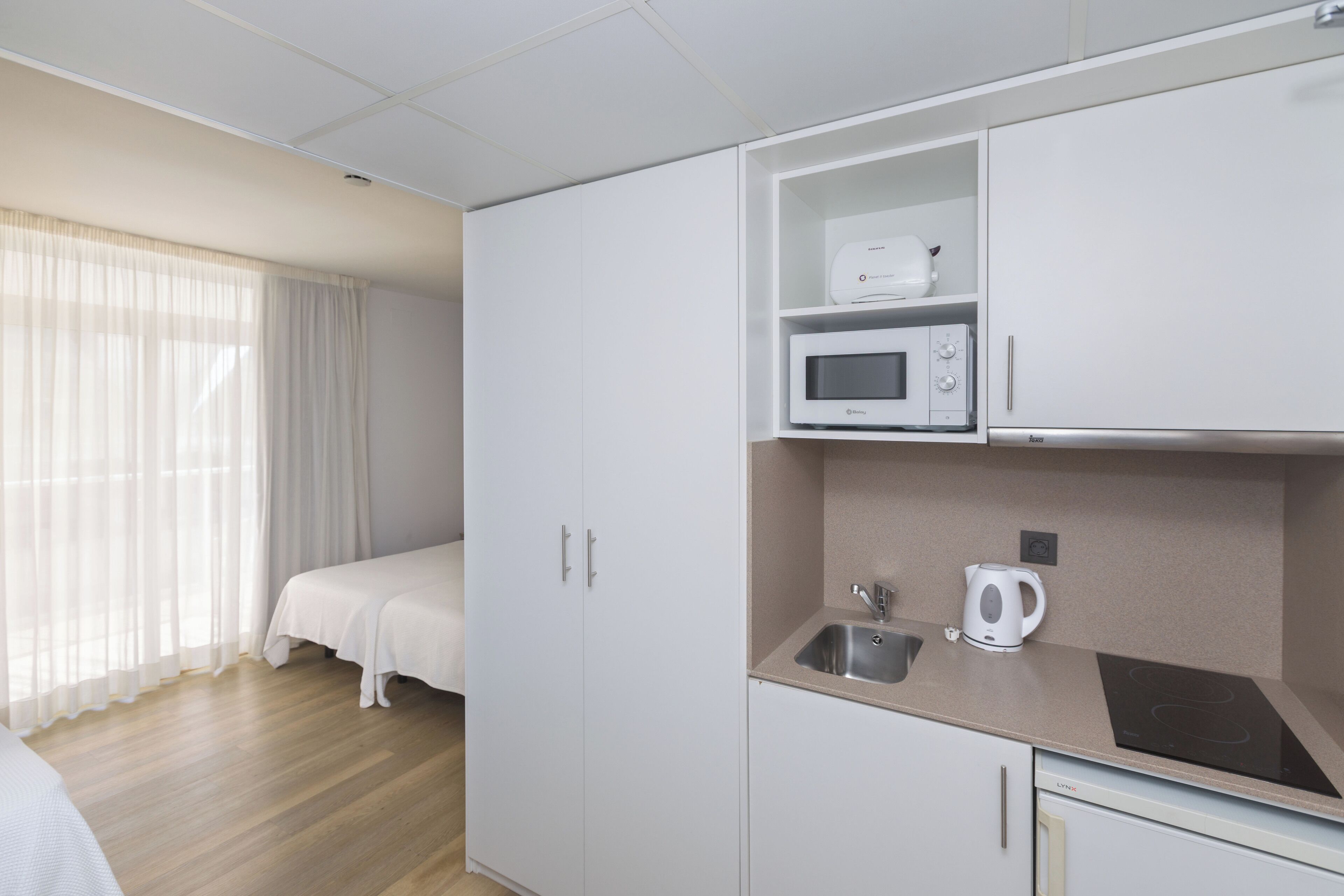 A compact hotel room with a kitchenette, a bed, and a window with curtains.