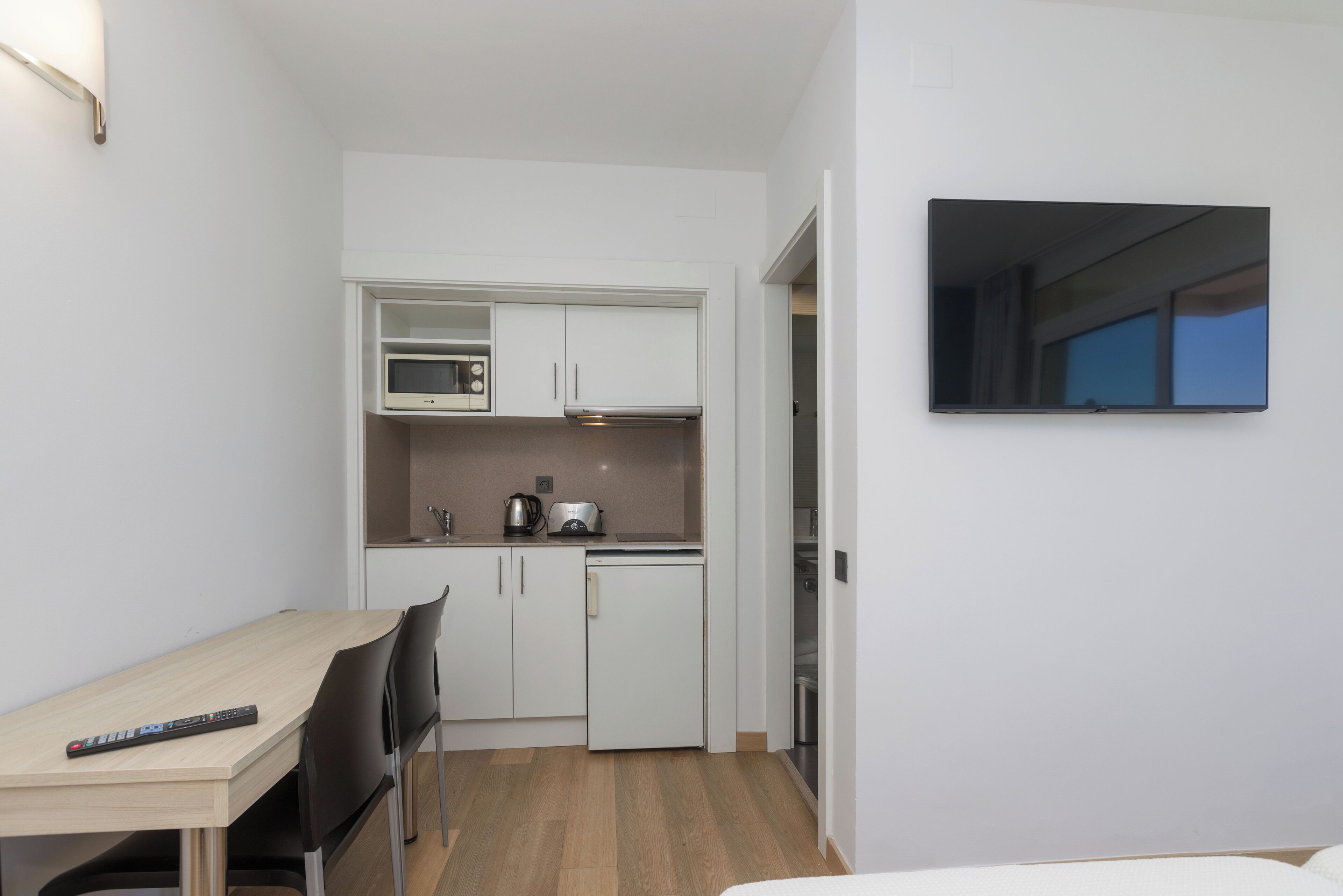 Cocina compacta con microondas, tostadora y refrigerador. Se observa una pequeña mesa de comedor con sillas y un televisor fijado a la pared.