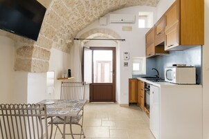 Una cocina compacta con piso de baldosas, una mesa de comedor con sillas, un televisor montado en la pared y armarios de madera.