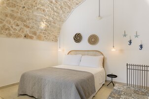 Un dormitorio con pared de piedra, una cama con colcha gris, dos platos decorativos colgados en la pared y una mesita con bandeja negra.