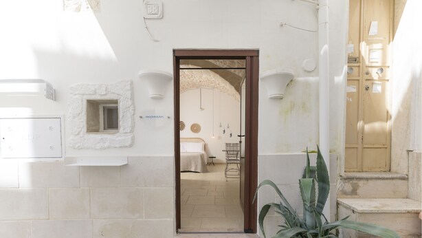 Een deuropening die leidt naar een kamer met een bed, een klein tafeltje en een stoel. Rechts van de deuropening staat een potplant.