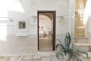 Un umbral da paso a una habitación con una cama, una mesita y una silla. A la derecha del umbral hay una planta en maceta.
