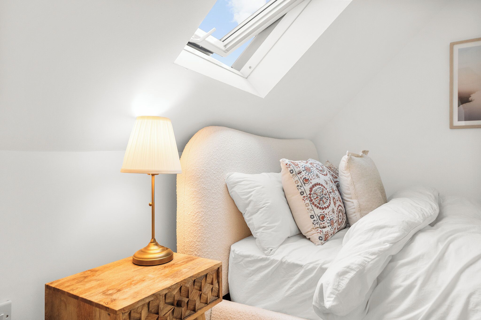 Een slaapkamer met dakraam, een bedlampje en een bed met witte beddengoed en opvallende kussens.