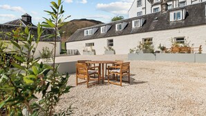 A courtyard with a gravel surface, a wooden table with chairs, and a building with multiple windows and a sloped roof. - Luxury 2 Bedroom Cottage, with private courtyard and garden. (Killin)