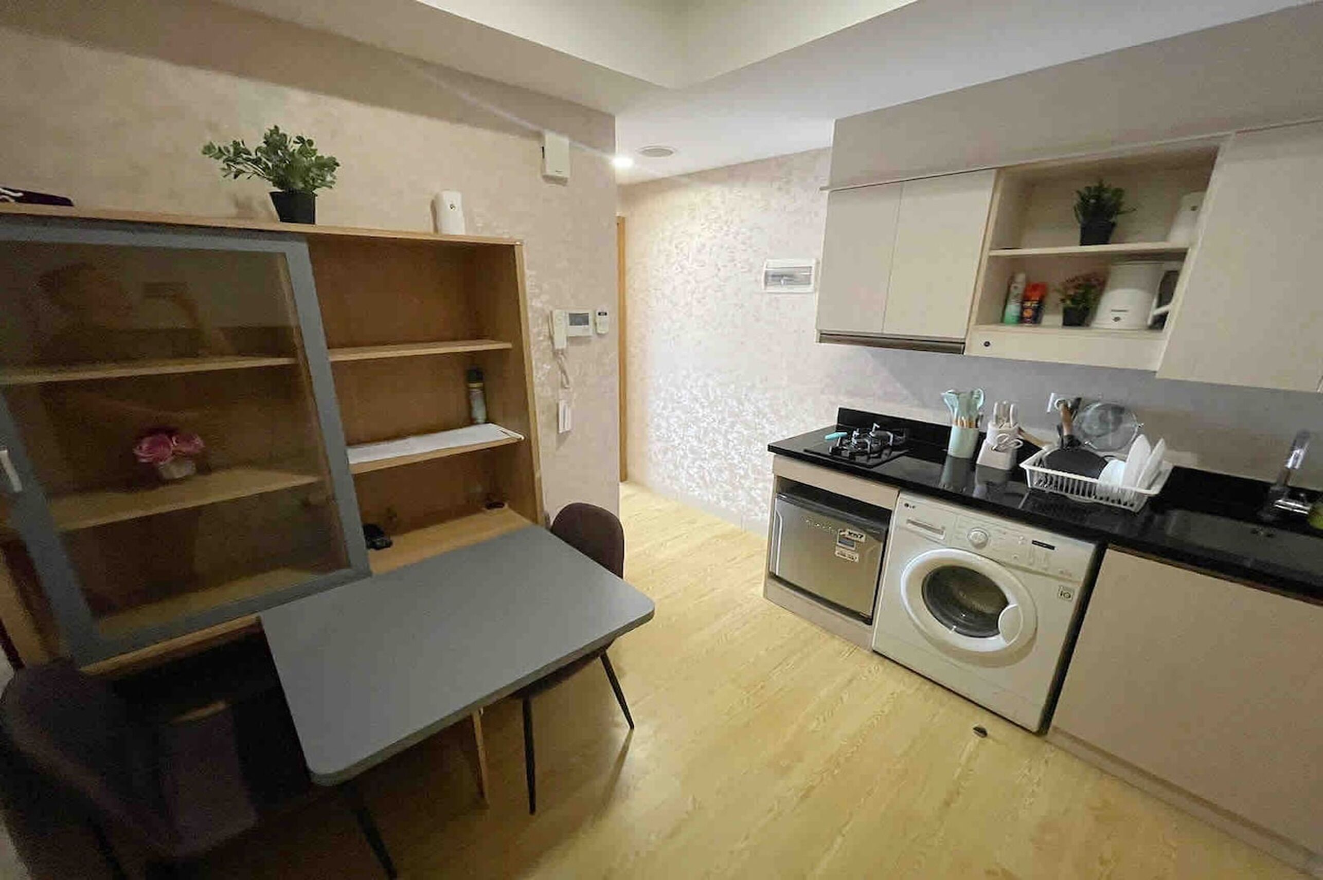 A compact kitchen with a washing machine, a dining table with chairs, and a built-in shelf with a potted plant.