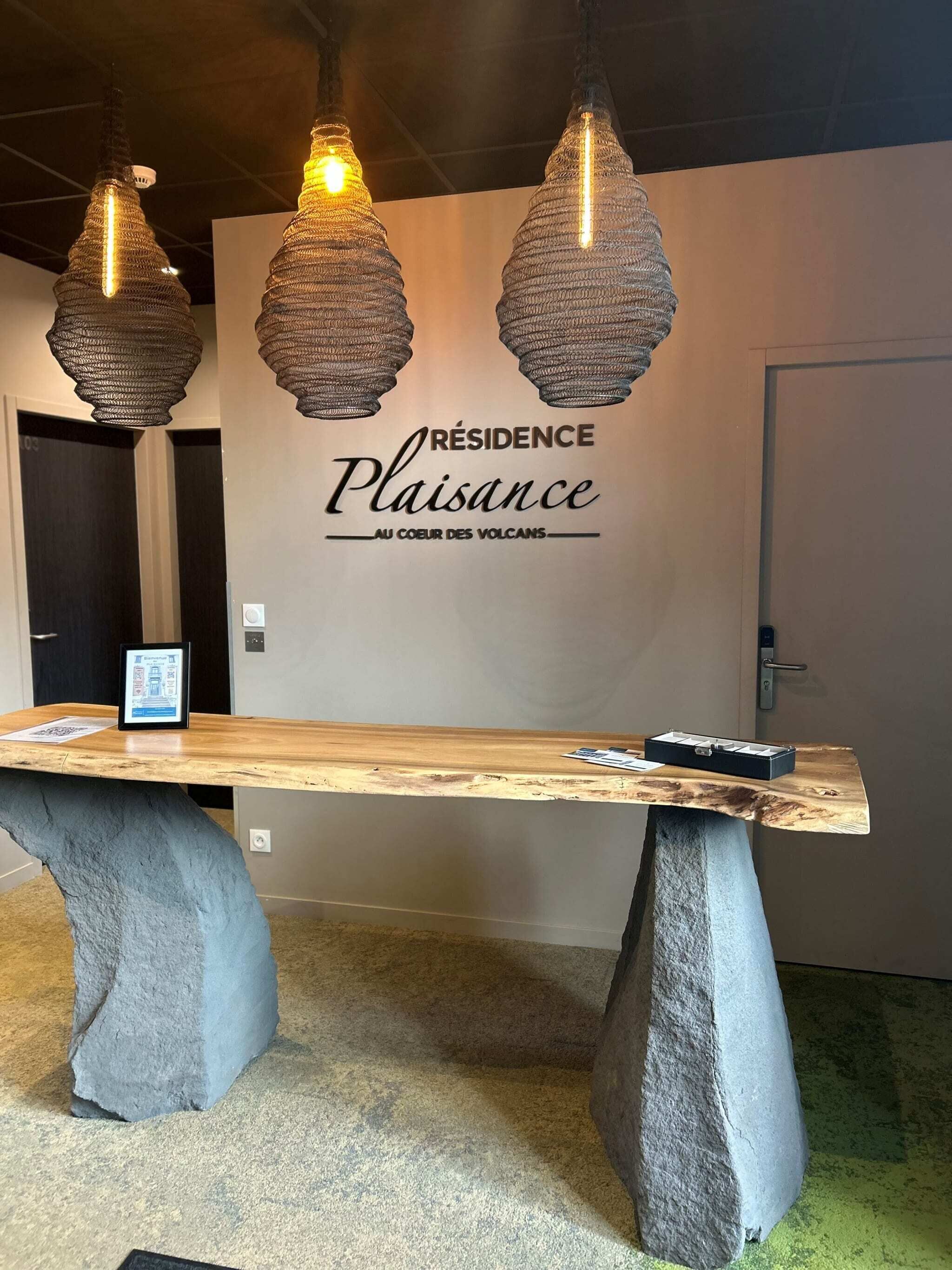 En receptionsdisk med en trätopp och socklar i sten, tre hängande lampor och en väggskylt med texten ”Résidence Plaisance Au Coeur Des Volcans”.
