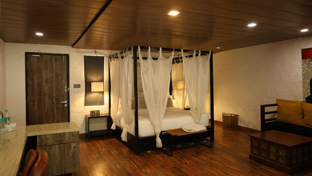 A bedroom with a four-poster bed, wooden flooring, a desk with a chair, a bedside table, and a sofa with cushions.