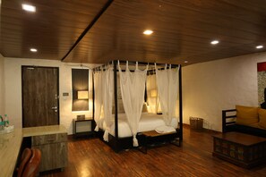 A bedroom with a four-poster bed, wooden flooring, a desk with a chair, a bedside table, and a sofa with cushions.