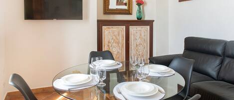 A dining area with a round glass table set for four, surrounded by black leather chairs. A flat-screen TV is mounted on the wall above the table.