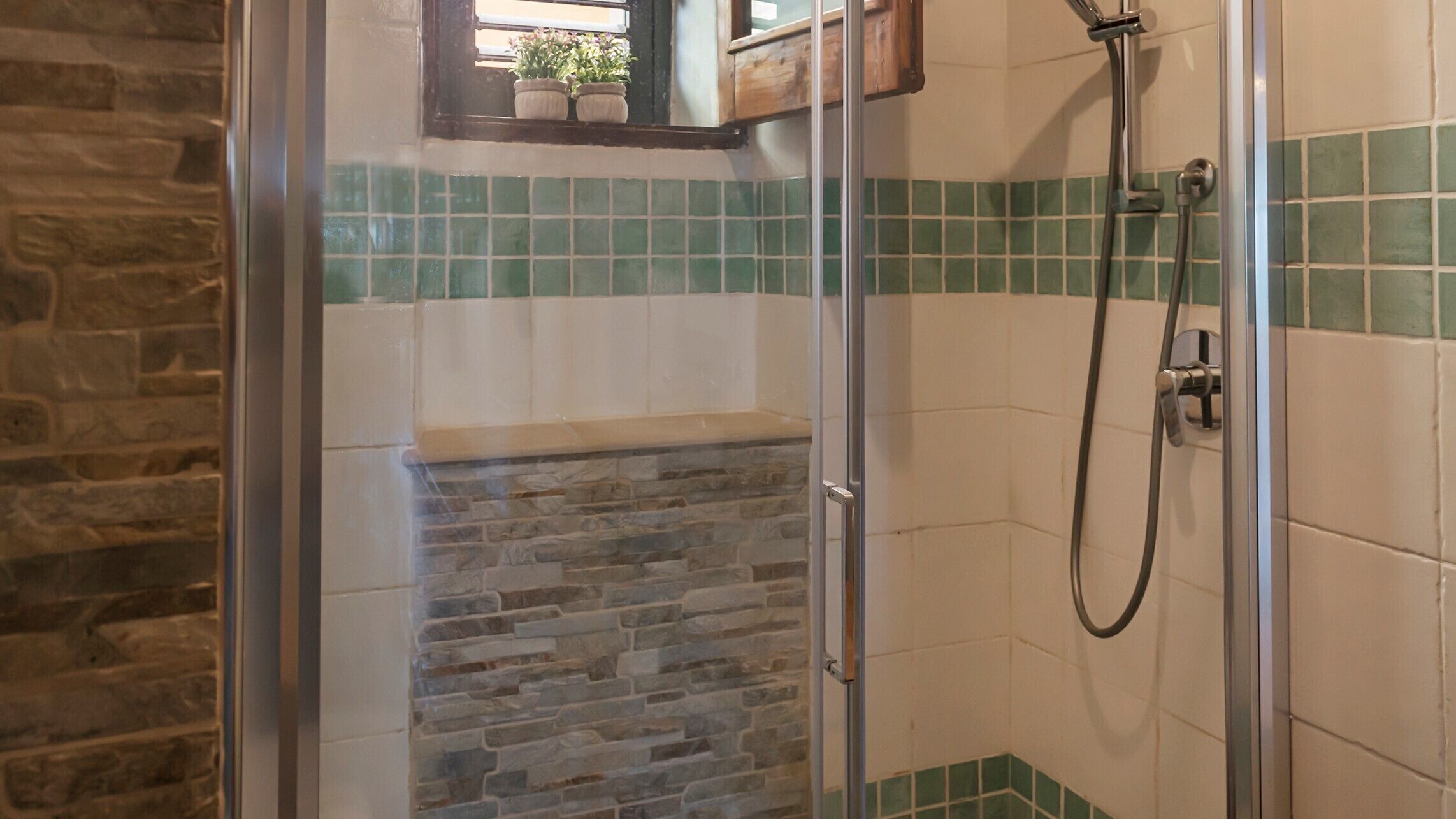 A shower area with a glass door, stone wall, and green-tiled walls.