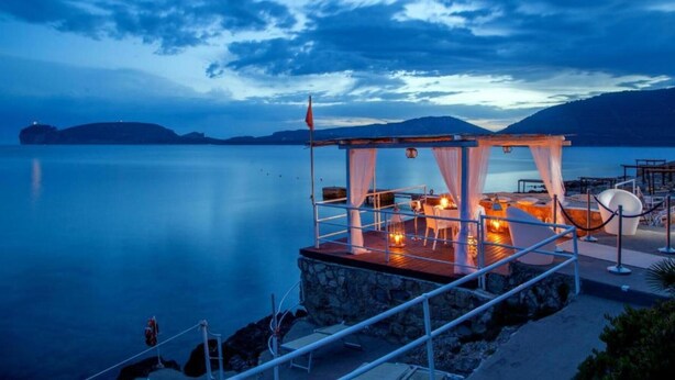 Une terrasse en bord de mer dotée d’un espace salon abrité, de murs en pierre et offrant une vue sur la mer calme et les montagnes lointaines.