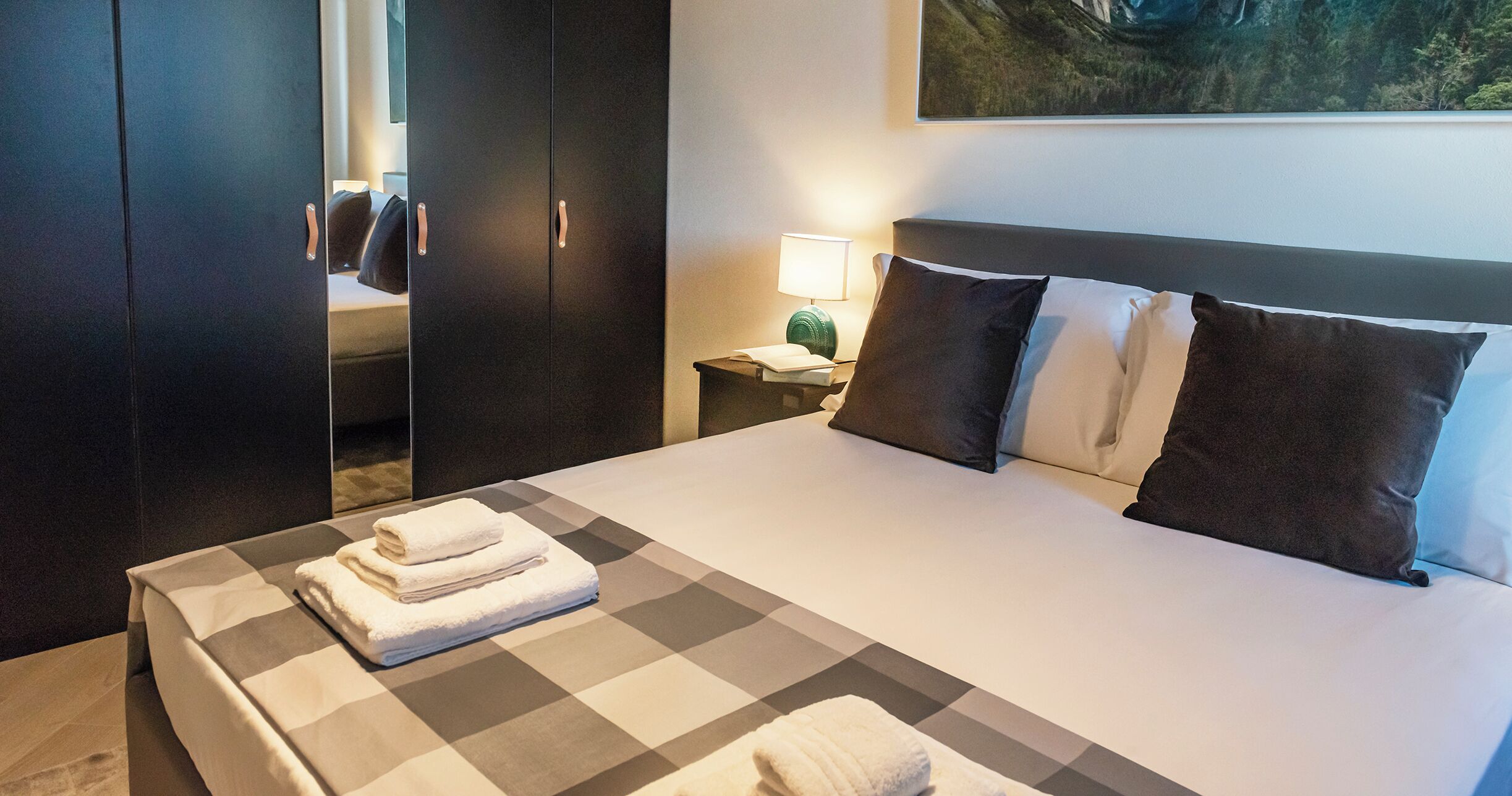 A hotel room with a large bed, a wardrobe, a nightstand with a lamp, and a painting on the wall.