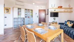 A dining area with a wooden table set for six, surrounded by wicker chairs. A sofa and a television are visible in the background.
