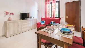 Een eetgedeelte met een houten tafel gedekt voor vier personen, een bank, een televisie op een kast, en een raam met rode gordijnen.