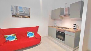 Uma cozinha compacta com fogão a gás, forno e uma pequena bancada. Um sofá vermelho com almofadas decorativas está posicionado em frente à cozinha.