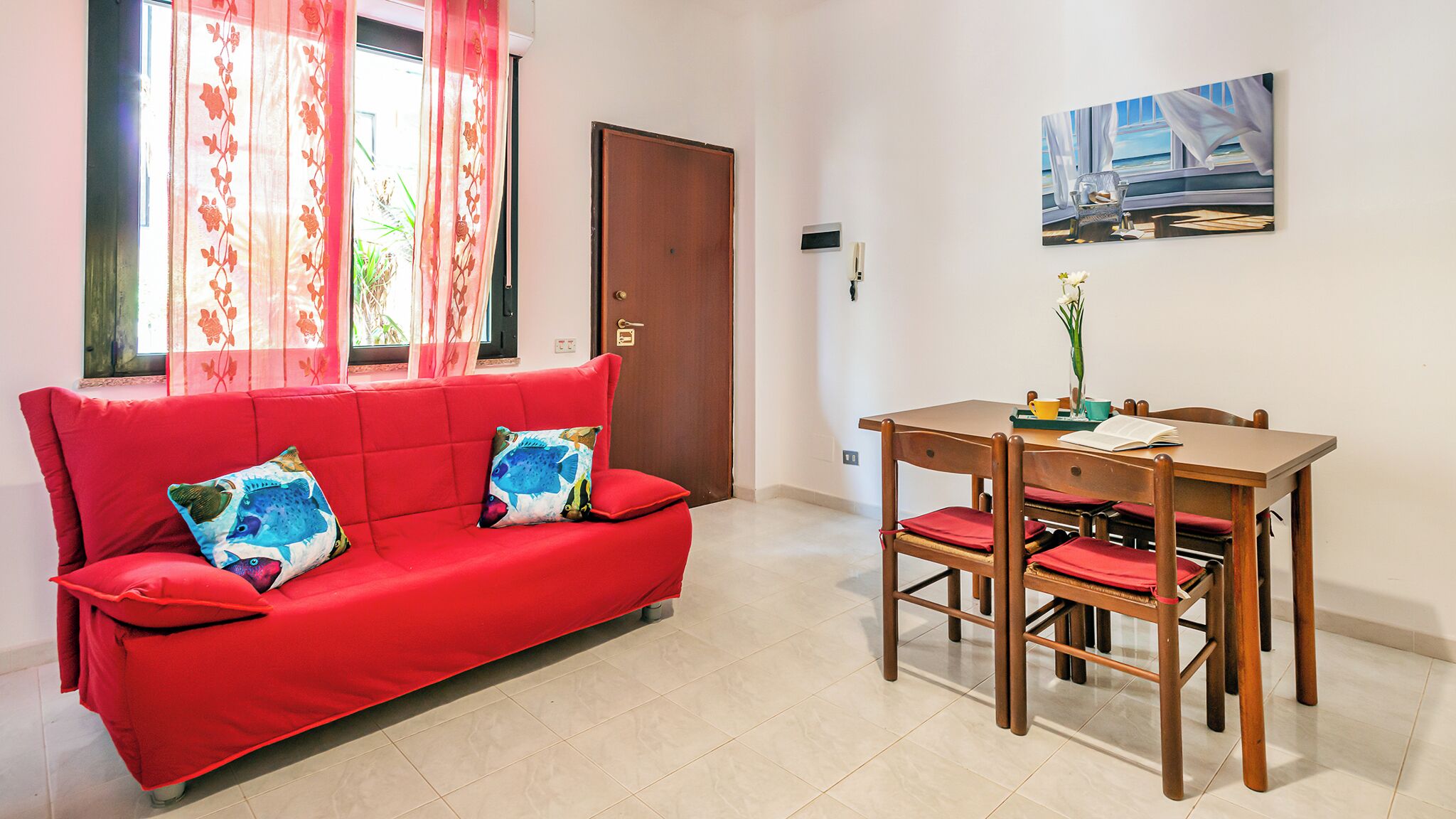 Um sofá vermelho com almofadas coloridas, uma mesa de jantar de madeira com cadeiras e um quadro na parede.