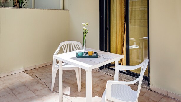 Een klein terras met een witte tafel en stoelen, een vaas met bloemen en een dienblad met een mok en een kopje.