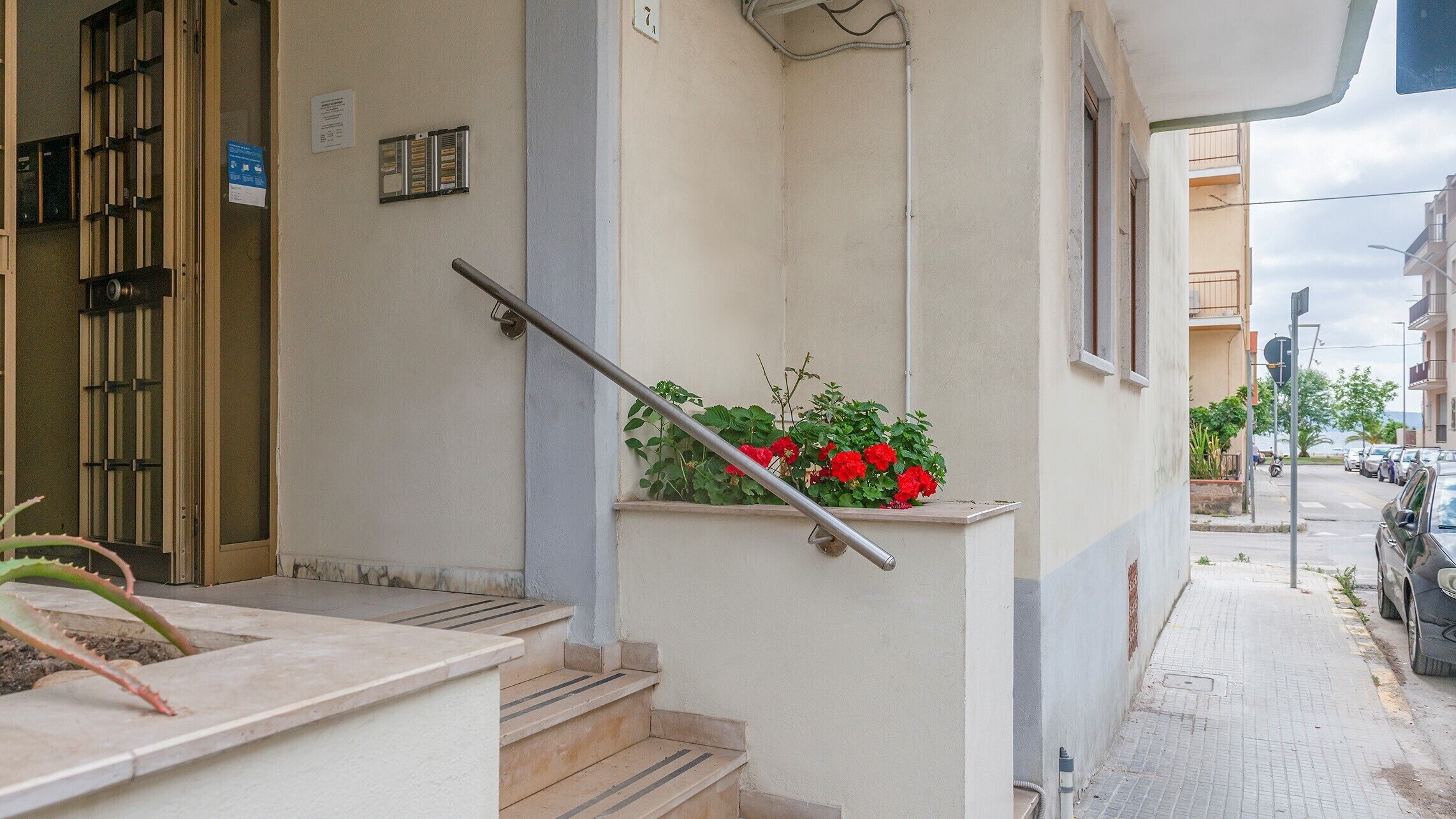 Edifício residencial com escadaria que dá para um patamar, portão de metal e um floreiro com flores vermelhas.