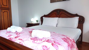Ein ordentlich bezogenes Bett mit floralem Bettbezug, zwei aufgerollte Handtücher und ein Nachttisch mit Lampe und einer kleinen Pflanze.
