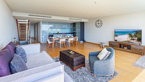 Un salon moderne avec un canapé, un fauteuil, une table basse et un téléviseur. Un escalier mène à un niveau supérieur, et on aperçoit une cuisine au fond.