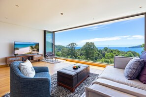 Una sala de estar moderna con un ventanal que ofrece una vista panorámica del océano y las colinas.