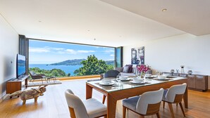 Un espace de restauration moderne avec une grande table en verre, des chaises en bois et une vue panoramique sur la mer et les collines.