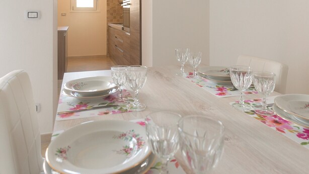 A dining table set with floral tablecloth, plates, and wine glasses in a room with a light-colored wooden floor and a view of a kitchen in the background.