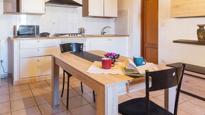 A compact kitchen with white cabinets, a microwave, a stove, and a sink. There's a dining table with chairs and a small potted plant. - Machin 3 - One Bedroom Apartment, Sleeps 2 (Alghero)