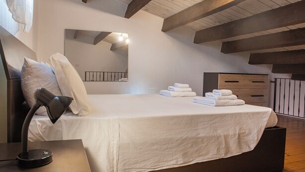 A neatly made bed with white linens and towels, a bedside lamp, a mirror, and a wooden dresser in a room with exposed beams on the ceiling. - Machin 3 - One Bedroom Apartment, Sleeps 2 (Alghero)