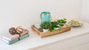 Ein Holzboden mit einer Stapel Bücher, einem gewebten Ball, einer Kokosnuss, einer türkisfarbenen Wasserflasche und einer Holztasse mit Pflanzen und einer Fruchtschale.