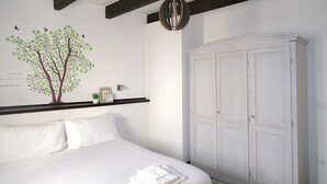 A bedroom with a bed, a white wardrobe, a wall-mounted shelf with decorative items, and a wall decal of a tree.