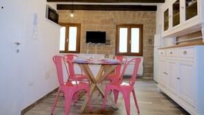 A dining area with a round table, pink chairs, and a wooden floor. There is a kitchen area with white cabinets and a TV mounted on the wall.