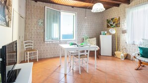Ein gemütliches Zimmer mit Essbereich, einem weißen Tisch und Stühlen. Es gibt ein Fenster mit Meerblick, eine weiße Kommode und eine Pendelleuchte.
