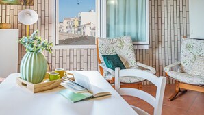 Ein Zimmer mit Blick auf eine Küstenstadt, ein Esstisch mit Büchern und einer Vase sowie zwei stilisierte Stühle.