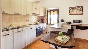 Una cocina con alacenas de madera clara, un lavamanos de acero inoxidable, un horno empotrado, un microondas y una mesa de madera con una bandeja de comida y una planta en maceta.