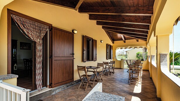 Un patio al aire libre con mesas y sillas de madera, una zona cubierta con suelo de baldosas y vistas a zonas verdes.