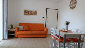 A living room with a sofa, dining table, chairs, a clock, and a door. - Prestigiosa - Two Bedroom Apartment, Sleeps 5 (Alghero)