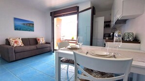 A dining area with a table set for two, a sofa with floral cushions, and a view of the sea through a glass door.
