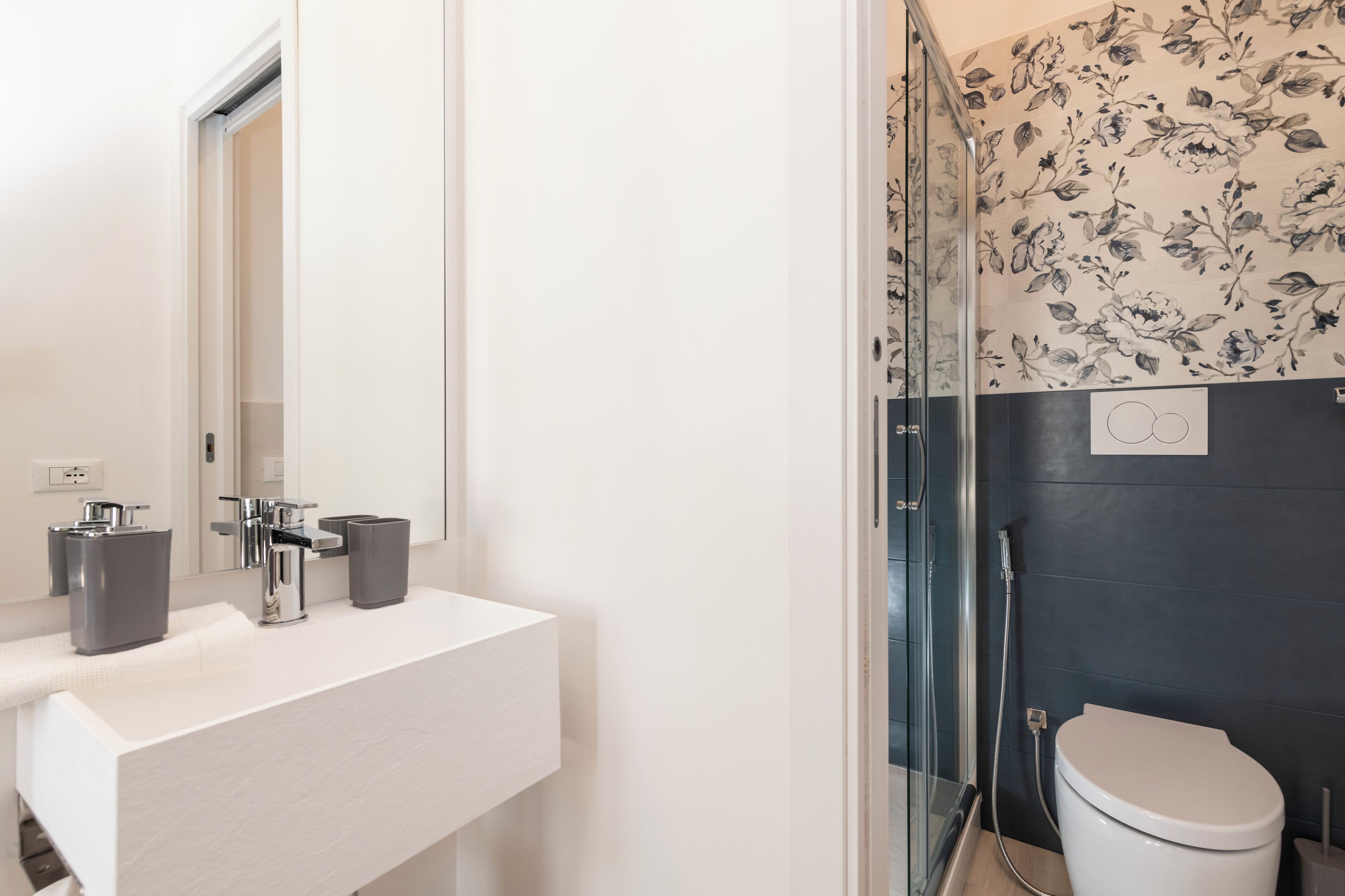 A modern bathroom with a white vanity, a glass shower enclosure, and a dark wall with floral wallpaper.