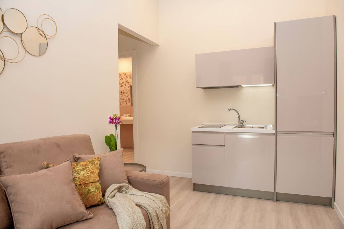 A modern kitchen with a built-in oven and a sink, a beige sofa with decorative pillows, and a wall-mounted mirror.