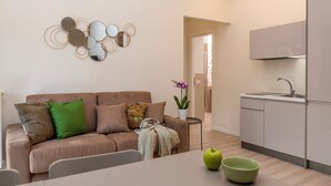 A modern living room with a beige sofa, a small side table with a potted plant, and a dining area with a table and chairs. A kitchen area is visible with cabinets and a sink. - Best Vatican Apartment (Rome)