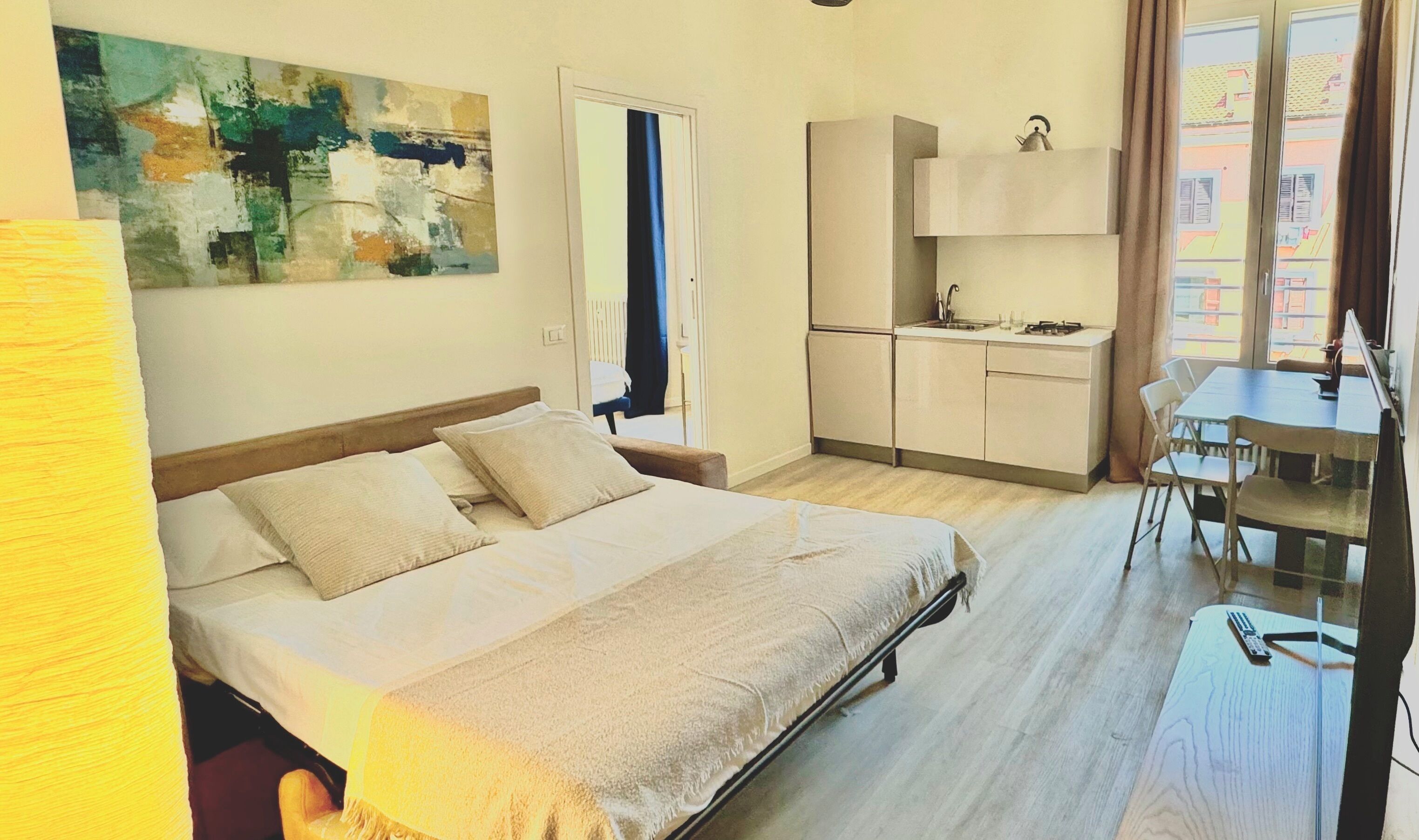 A hotel room with a bed, a small kitchenette, a dining table, and a window with curtains.