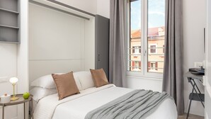 A bedroom with a bed, bedside table, lamp, and a view of buildings through a window. - Best Vatican Apartment (Rome)
