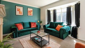 Un salon moderne aux murs de couleur turquoise, avec deux canapés verts ornés de coussins rouges et orange, une table basse en verre et une grande fenêtre agrémentée de rideaux noirs.