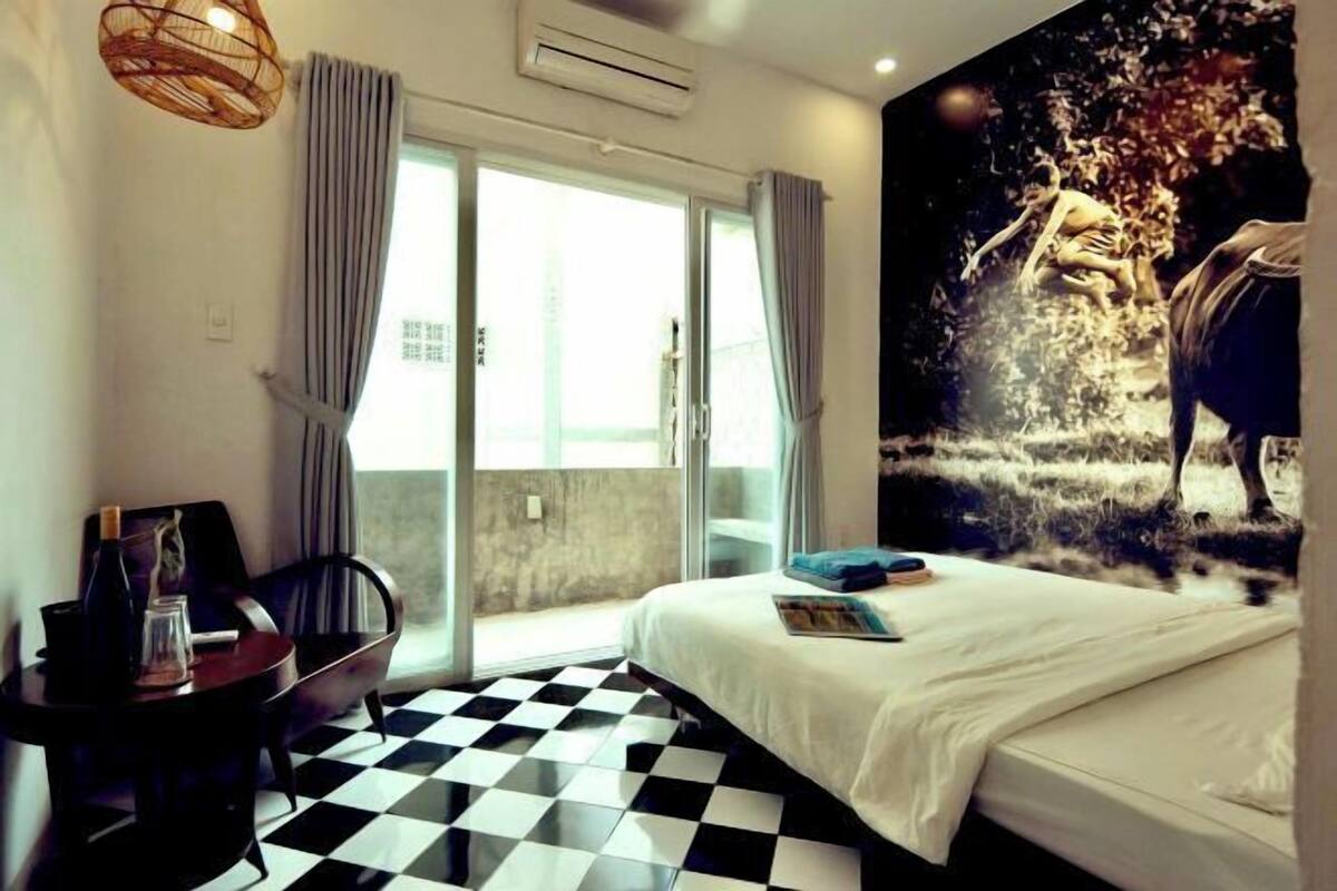 A hotel room with a checkered floor, a bed with white bedding, a large window with curtains, and a wall mural.