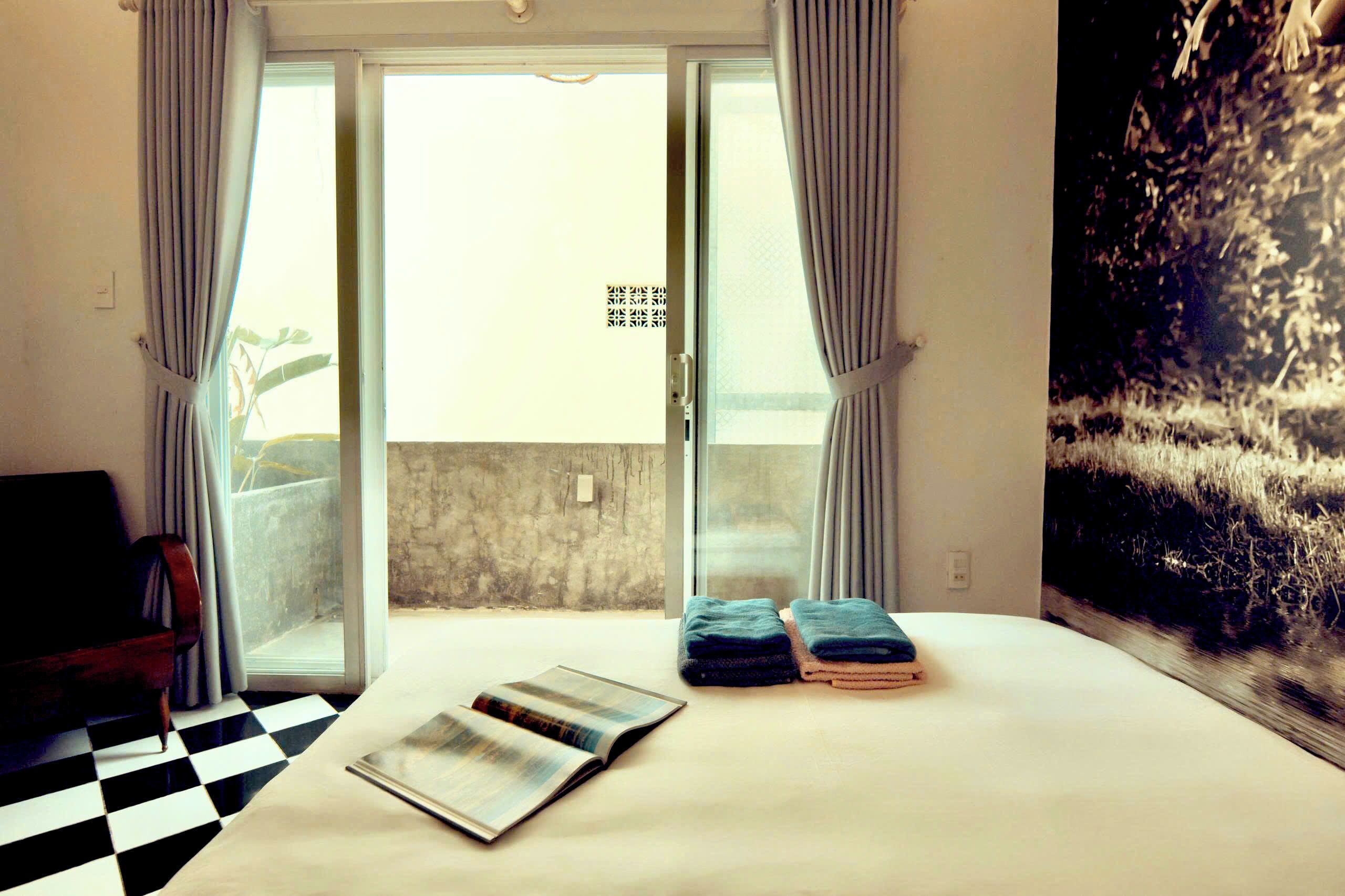 A hotel room with a checkered floor, a bed with folded towels, a magazine on the bed, and a view of a balcony through the door.