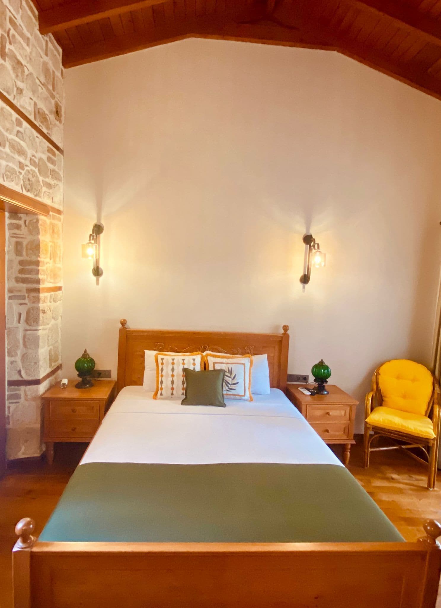 A bedroom with a wooden bed, two bedside tables, a nightstand, a lamp, a chair, and a stone wall.