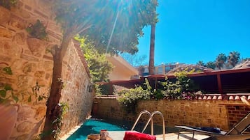 Zona junto a la piscina con una pared de piedra, un árbol y un cielo azul despejado.