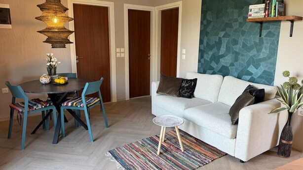 A modern living room with a white sofa, teal chairs, a dining table with a vase of flowers, a hanging light fixture, and a geometric-patterned accent wall. - Villa Feronia Samos (Mesokampos)