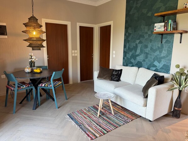 A modern living room with a white sofa, teal chairs, a dining table with a vase of flowers, a hanging light fixture, and a geometric-patterned accent wall.